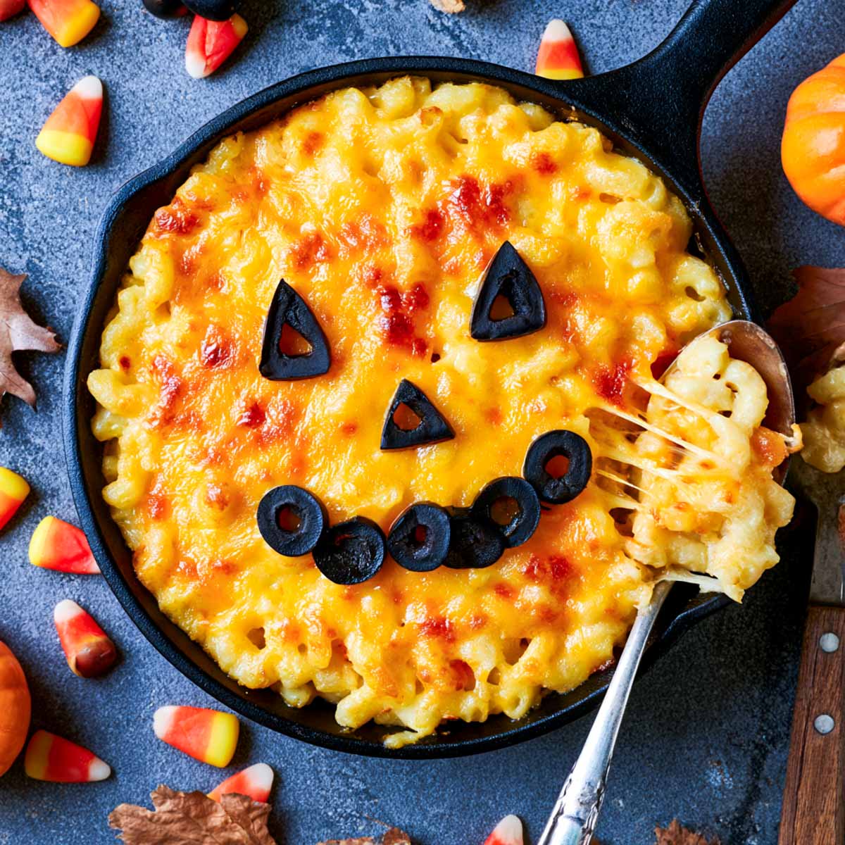 Skillet of Halloween mac and cheese with a jack-o-lantern face made from black olive pieces on the melted cheese.