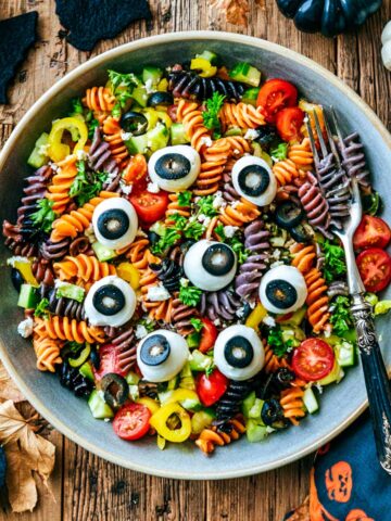 Overhead view of Halloween pasta salad with tri-color rotini, mozzarella eyeballs, cherry tomatoes, cucumber, peppers, olives, feta, and parsley in a gray bowl.