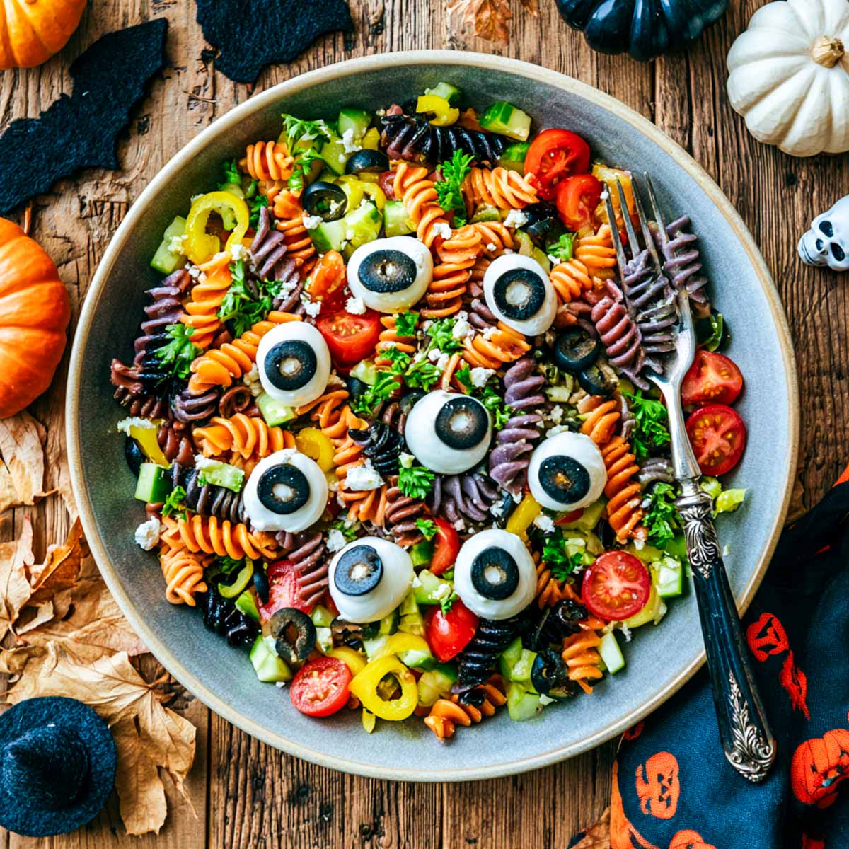 Overhead view of Halloween pasta salad with tri-color rotini, mozzarella eyeballs, cherry tomatoes, cucumber, peppers, olives, feta, and parsley in a gray bowl.