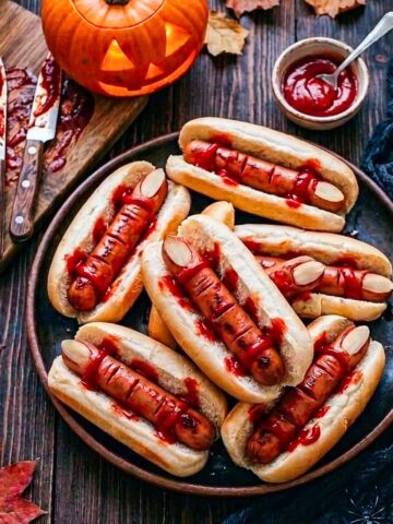 Close up of Halloween hot dog fingers with almond fingernails and ketchup served in buns on a dark platter.