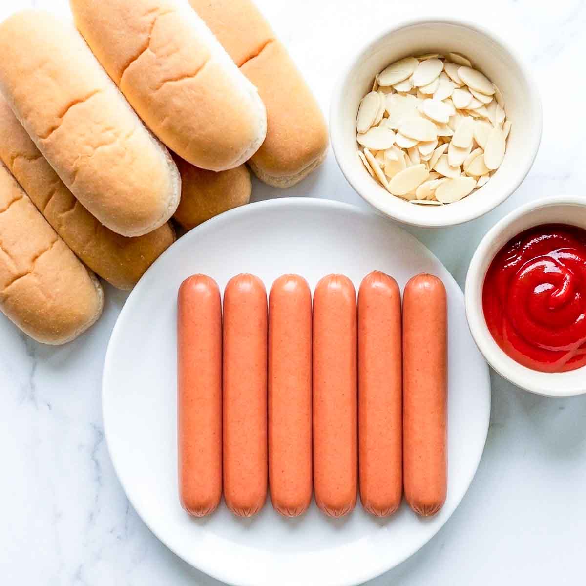 Hot dogs, buns, sliced almonds and ketchup laid out on a marble counter.