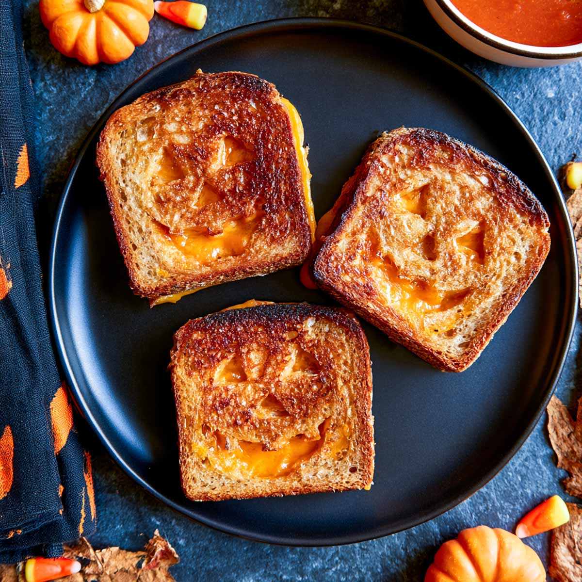 Three jack-o-lantern grilled cheese sandwiches with faces cut into the toasted bread showing melted orange cheese.
