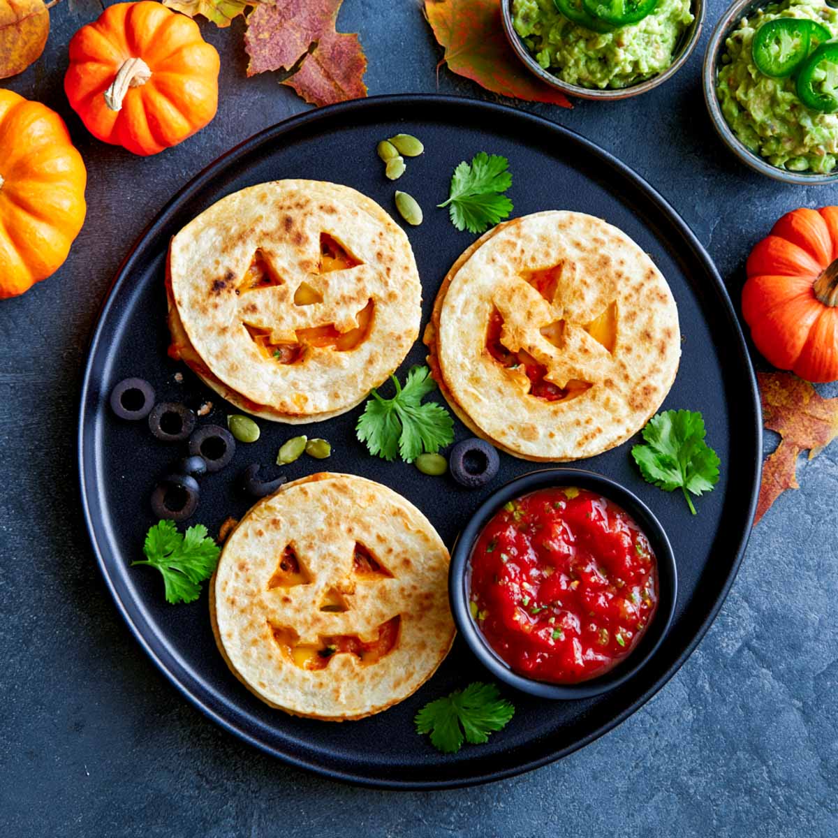 Three jack-o-lantern quesadillas with faces cut into the top tortilla showing melted orange cheese, with salsa and guacamole.