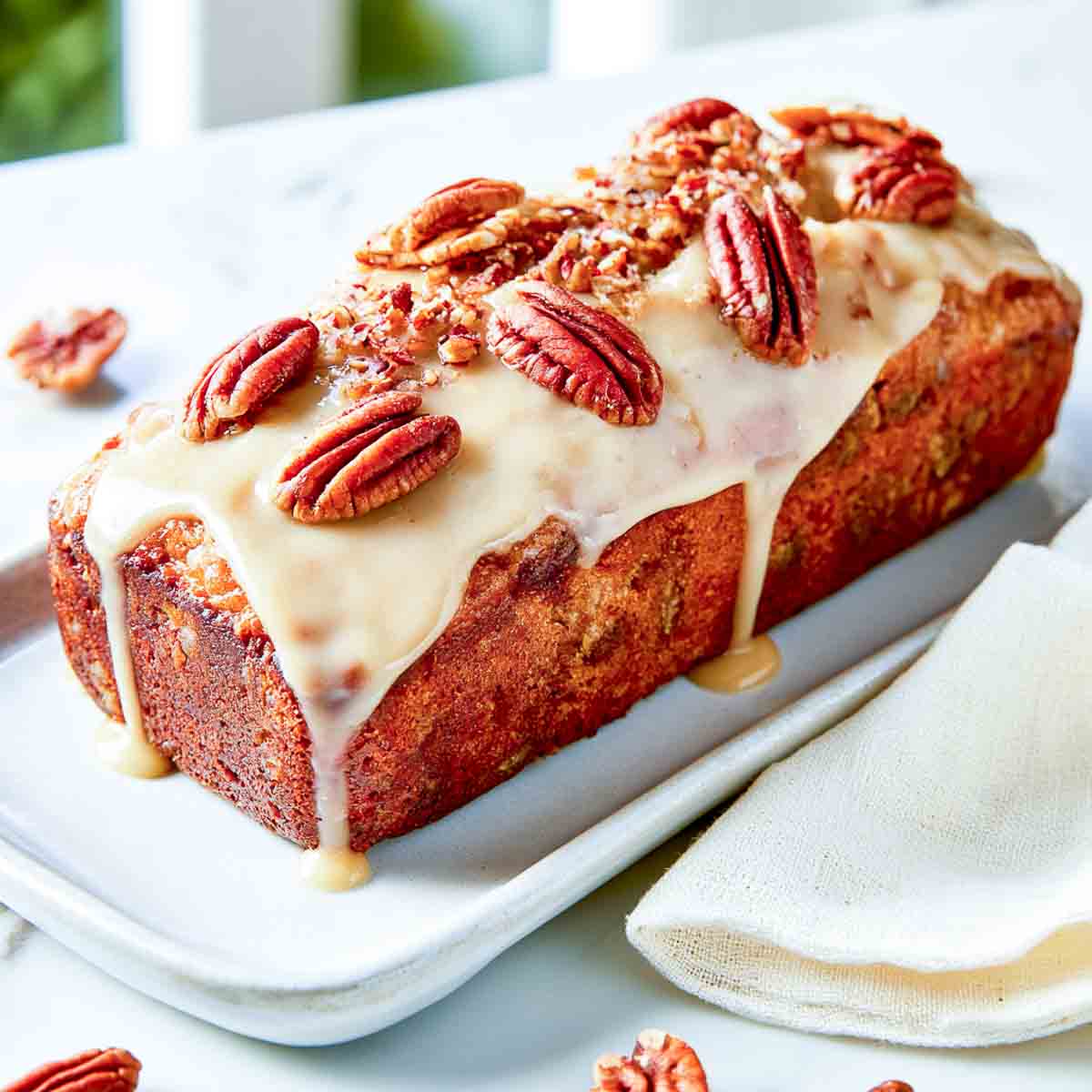 Maple pecan loaf cake topped with creamy maple glaze and whole pecans, arranged on a white rectangular serving plate.