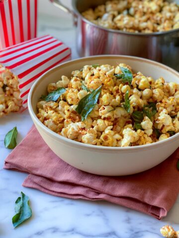 Bowl of masala popcorn tinted yellow with turmeric and studded with crisp curry leaves, beside striped popcorn cartons and a steel pot.