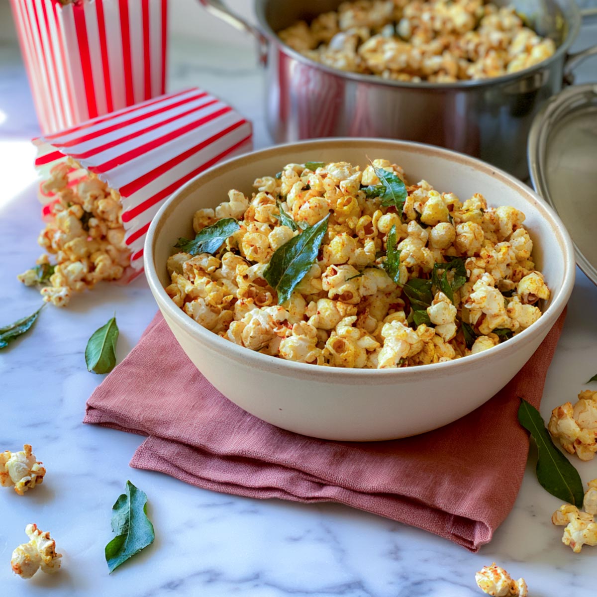 Bowl of masala popcorn tinted yellow with turmeric and studded with crisp curry leaves, beside striped popcorn cartons and a steel pot.