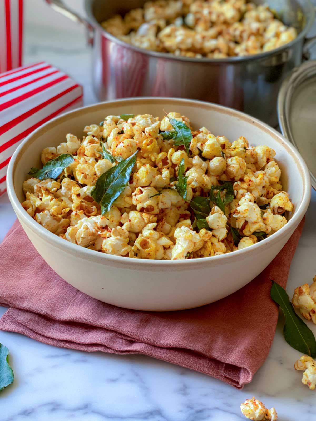Close-up of golden masala popcorn with crisp curry leaves in a cream ceramic bowl, styled on a pink linen napkin over white marble surface.