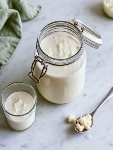Homemade milk kefir in a swing-top glass jar with kefir grains on the surface, a small glass of kefir, and a spoonful of milk kefir grains on white marble.