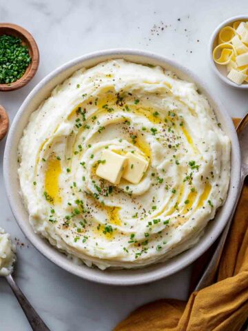 Creamy make-ahead mashed potatoes topped with melted butter, black pepper, and fresh chives in a white serving bowl, with extra butter, chives, and seasonings on a white marble surface.
