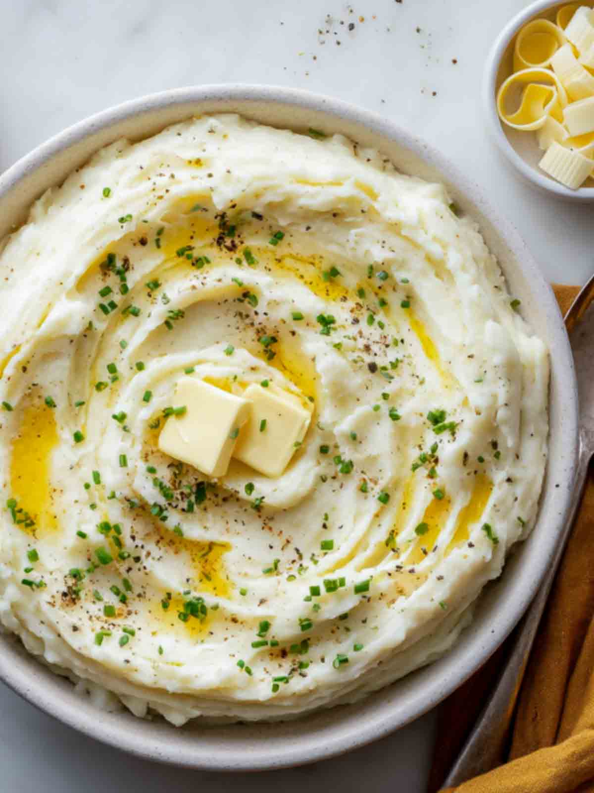 Close-up of creamy make-ahead mashed potatoes topped with melted butter, fresh chives, and black pepper in a white serving bowl.