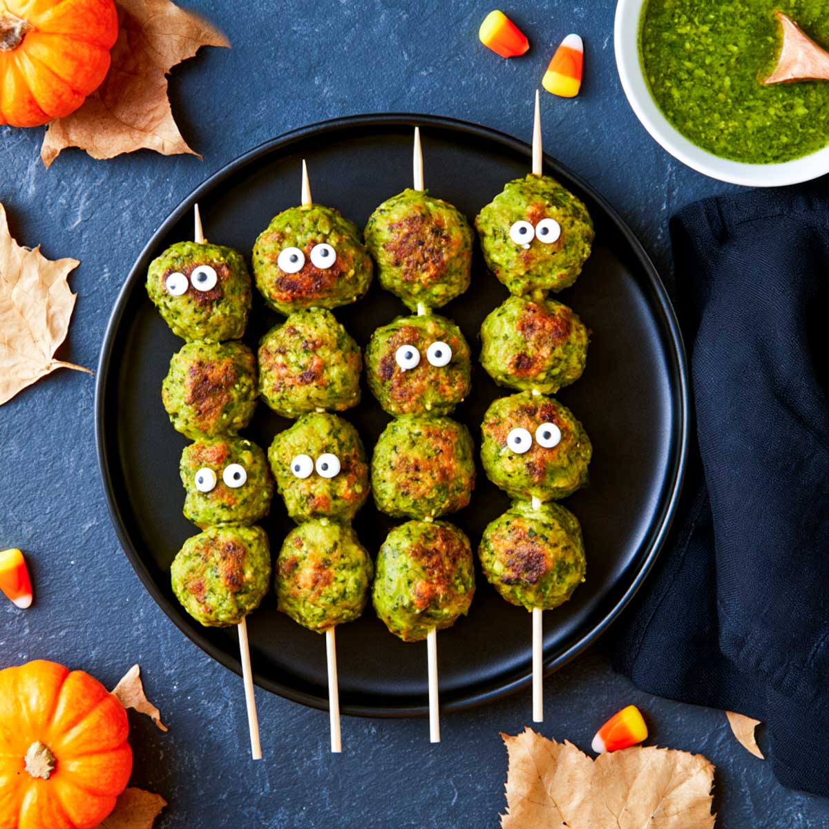 Platter of green pesto-coated monster meatballs on skewers with candy eyes, served with a bowl of pesto.