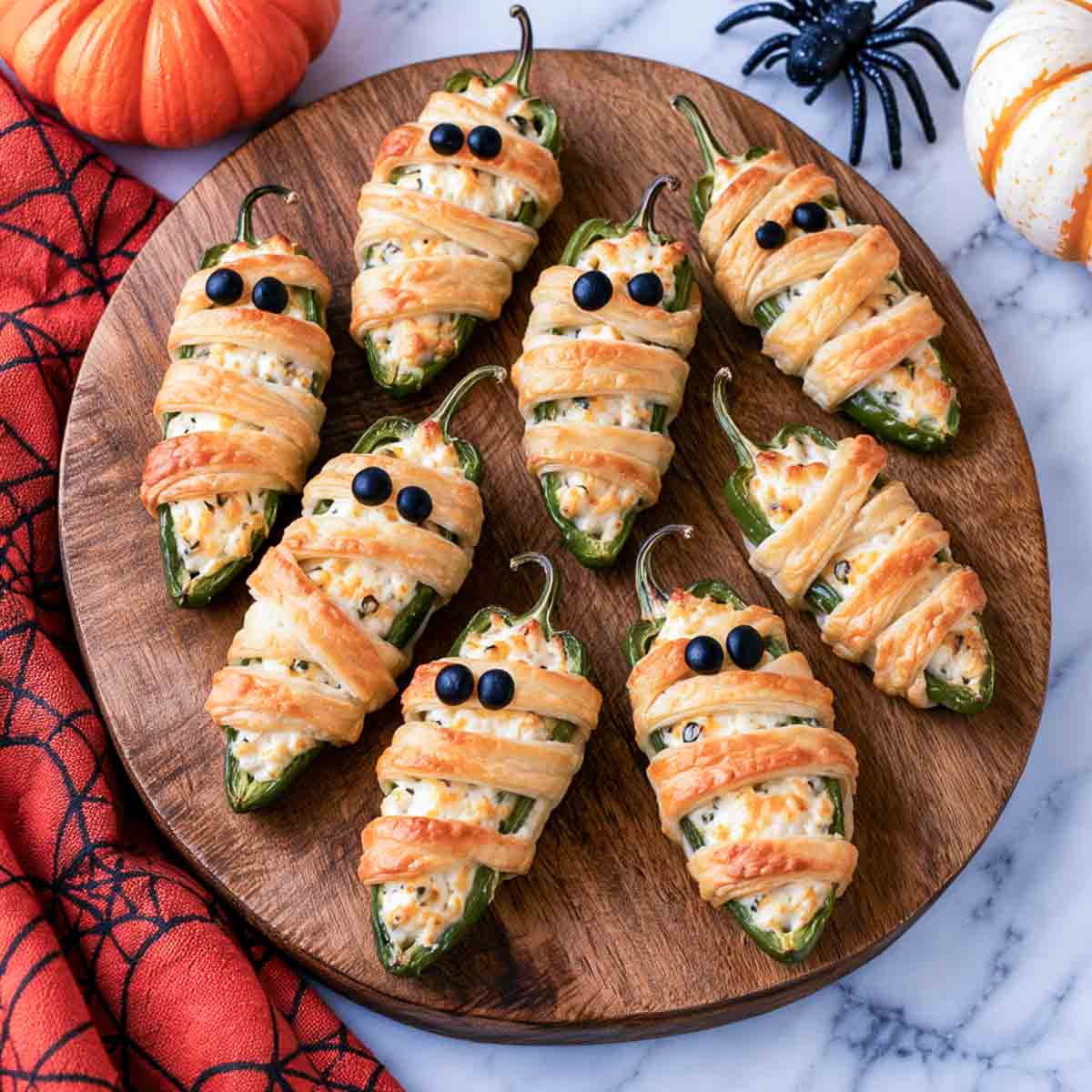 Mummy jalapeno poppers wrapped in pastry strips with black olive eyes on a round wood board.