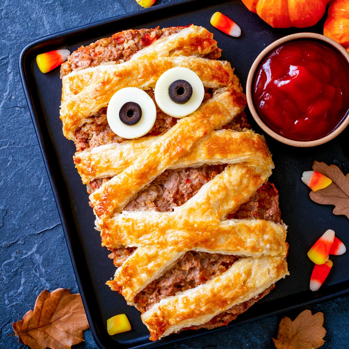 Mummy meatloaf wrapped in golden pastry strips with mozzarella and olive eyes, served with a bowl of ketchup.