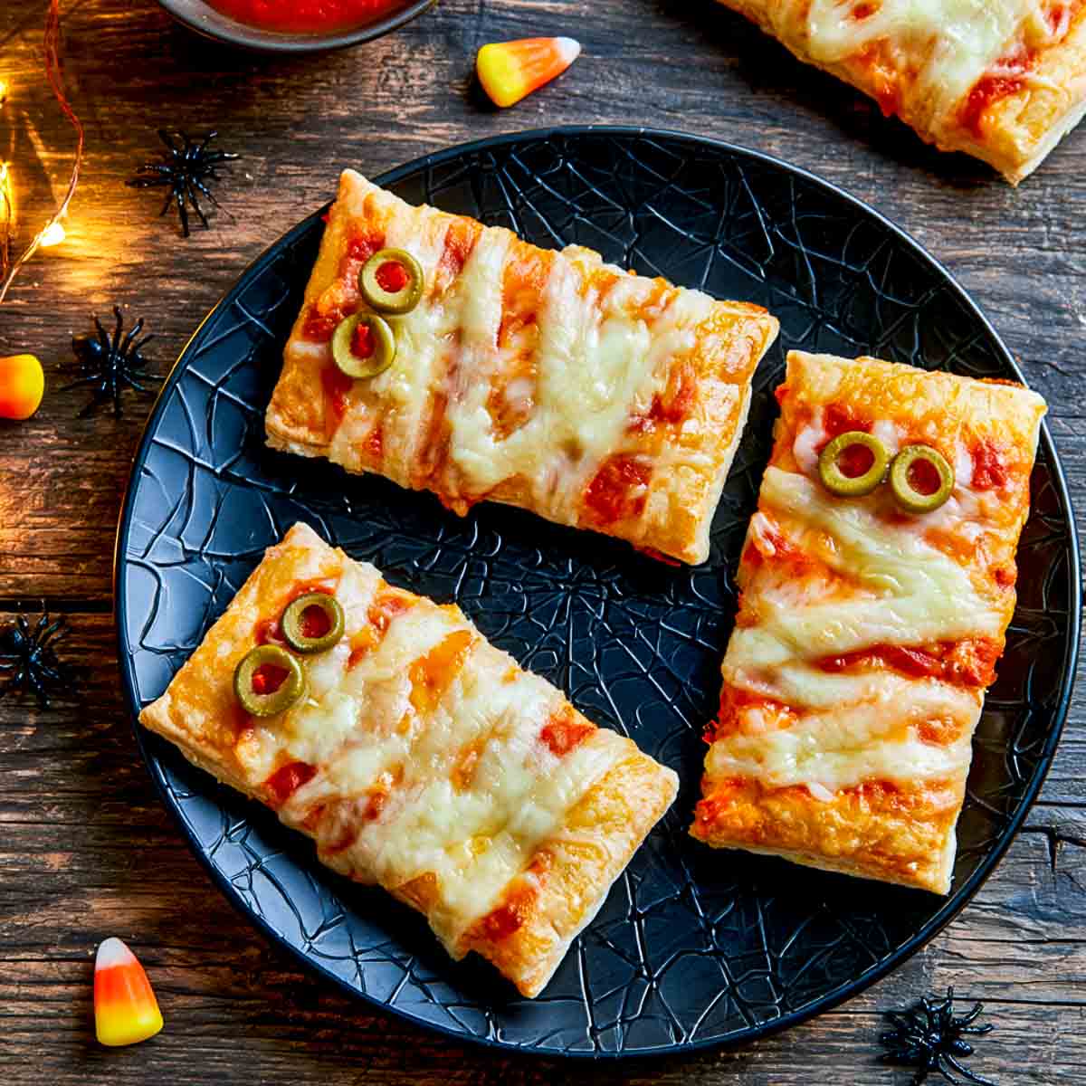 Close-up of three puff pastry mummy pizza bites showing thin mozzarella strips and green olive eyes.