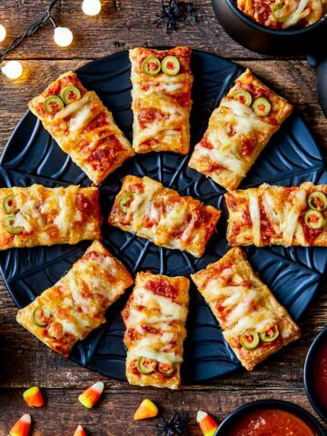 Overhead view of homemade mummy pizza bites arranged on a black Halloween plate, surrounded by candy corn, plastic spiders, and bowls of pizza sauce.