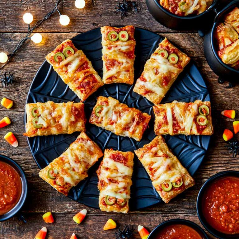 Overhead view of homemade mummy pizza bites arranged on a black Halloween plate, surrounded by candy corn, plastic spiders, and bowls of pizza sauce.
