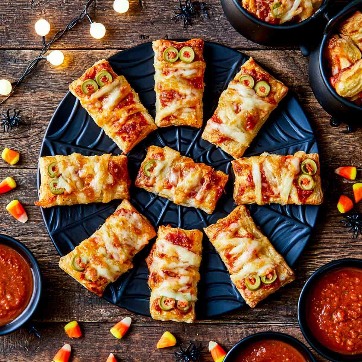 Overhead view of homemade mummy pizza bites arranged on a black Halloween plate, surrounded by candy corn, plastic spiders, and bowls of pizza sauce.