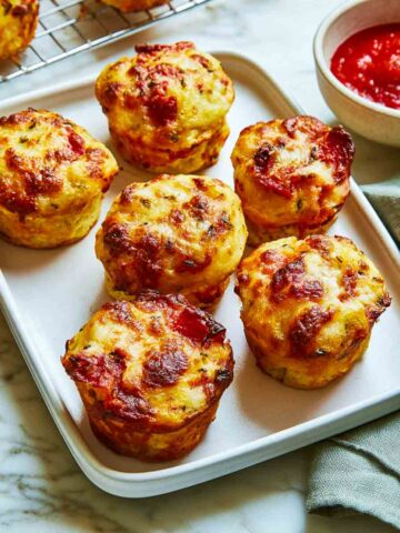 Six golden pizza muffins with browned cheese and pepperoni on a white tray beside a bowl of pizza sauce.