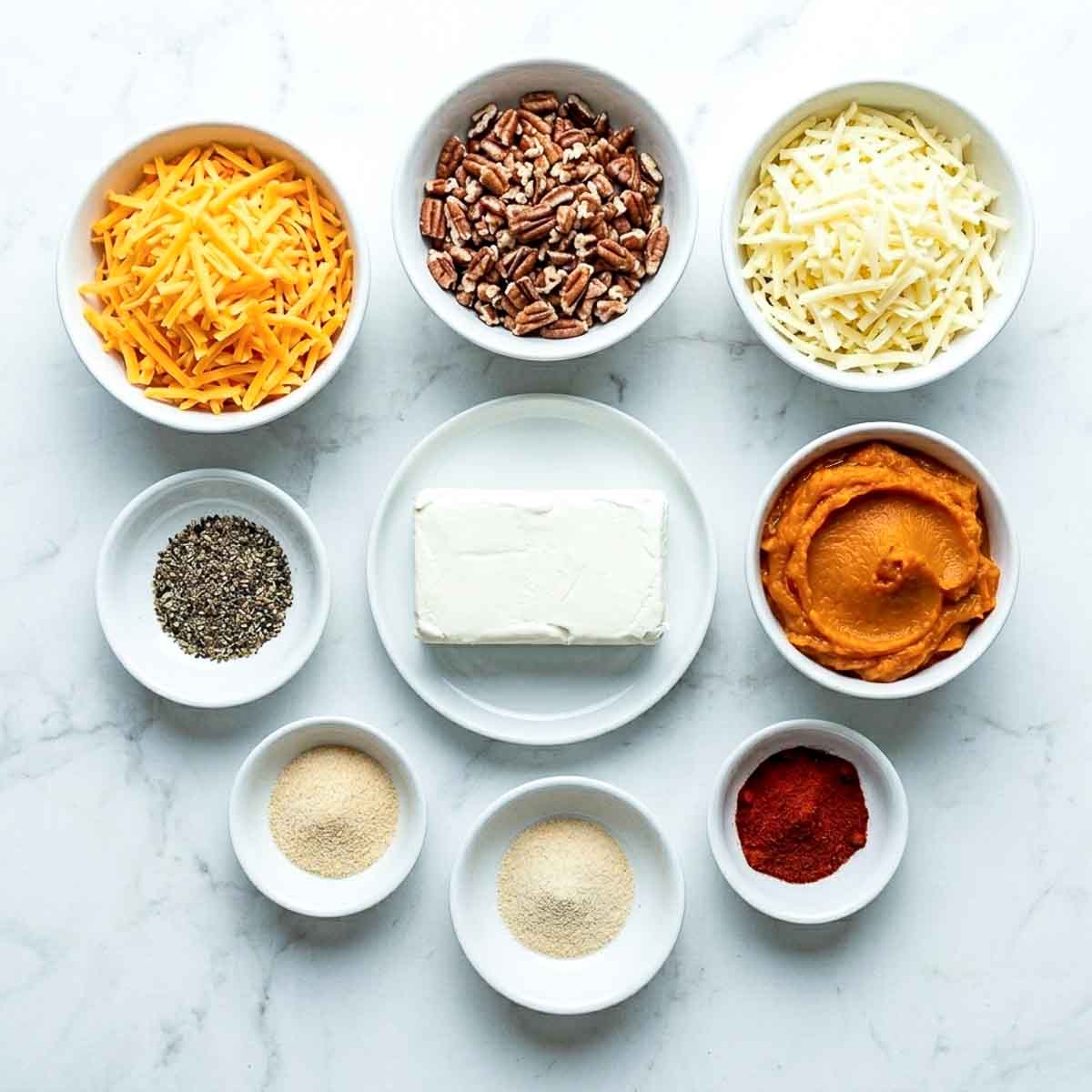 Bowls of shredded cheddar, shredded Monterey Jack, cream cheese, pumpkin puree, chopped pecans and spices arranged for a pumpkin cheese ball recipe.