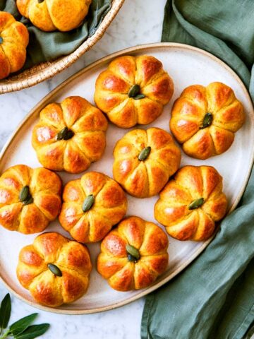 Nine golden pumpkin-shaped dinner rolls on a cream oval platter with a pepita in each center, green linen and butter alongside.