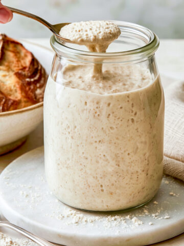 A spoonful of bubbly sourdough starter being lifted from a glass jar, with a loaf of sourdough bread and a linen napkin in the background.