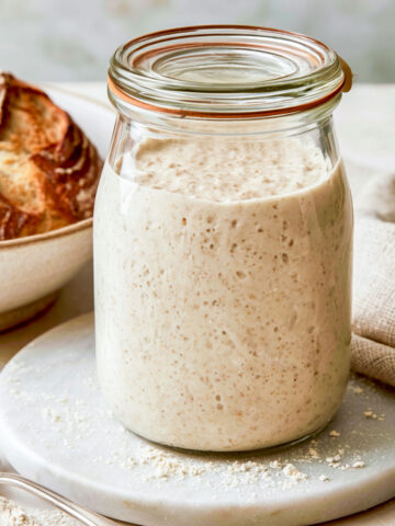 A jar of bubbly sourdough starter on a white marble surface beside a loaf of crusty sourdough bread and a linen napkin.