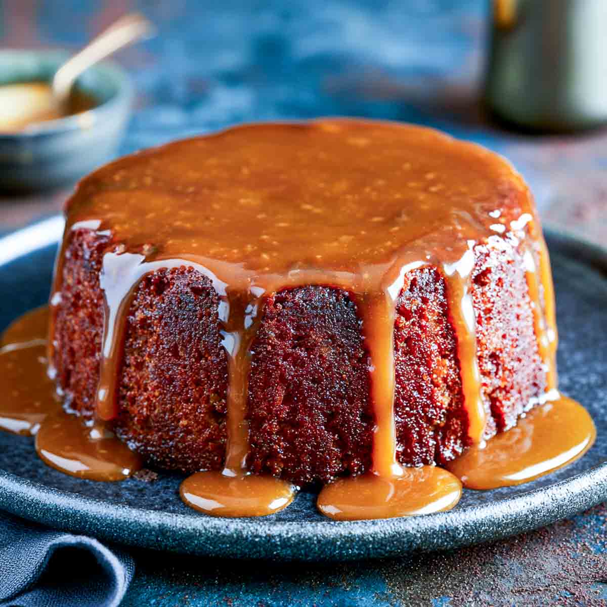 Sticky toffee cake covered in glossy caramel sauce that drips down the sides and pools around the base on a dark serving plate.