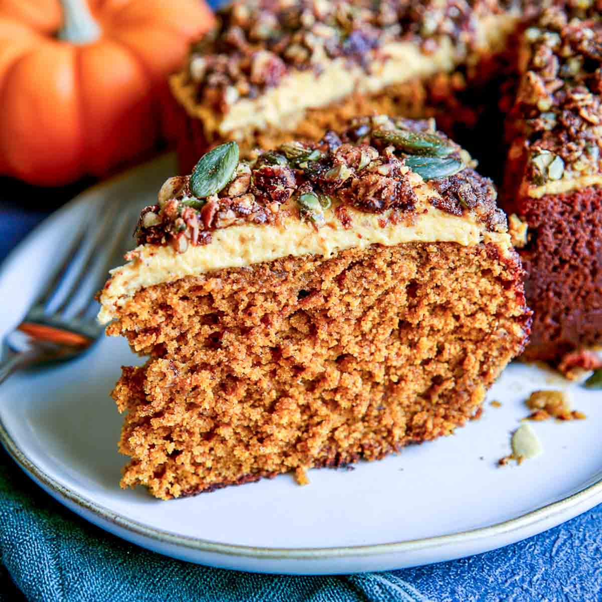 Slice of vegan pumpkin spice cake topped with creamy frosting, chopped nuts, and pumpkin seeds, with additional cake pieces in the background.