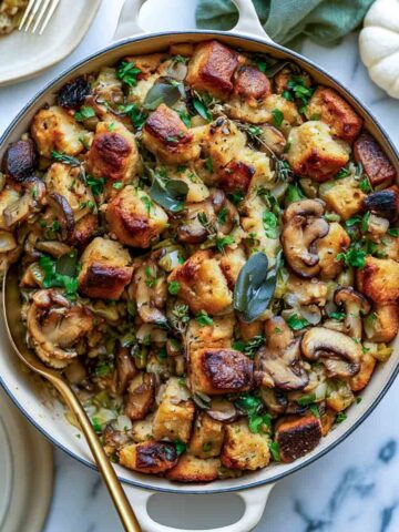 Top-down view of vegan sourdough stuffing with golden toasted bread cubes, browned mushrooms, celery, onions, parsley, sage, and thyme in a cream-colored Dutch oven on white marble.