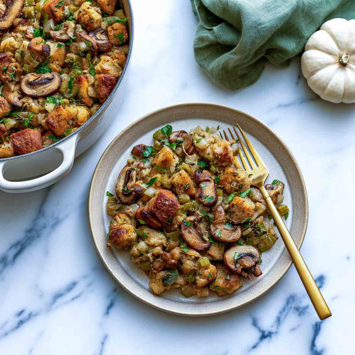 A serving of vegan sourdough stuffing with mushrooms and celery on a cream plate with a gold fork, the Dutch oven beside it.