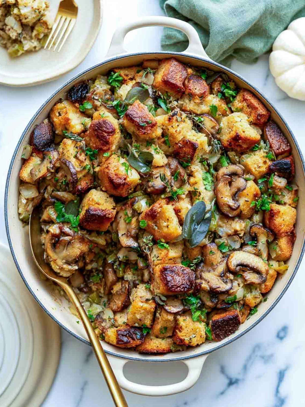 Mushroom sourdough stuffing in a cream Dutch oven with crisp bread cubes, sage leaves, and a plated serving beside a white pumpkin.