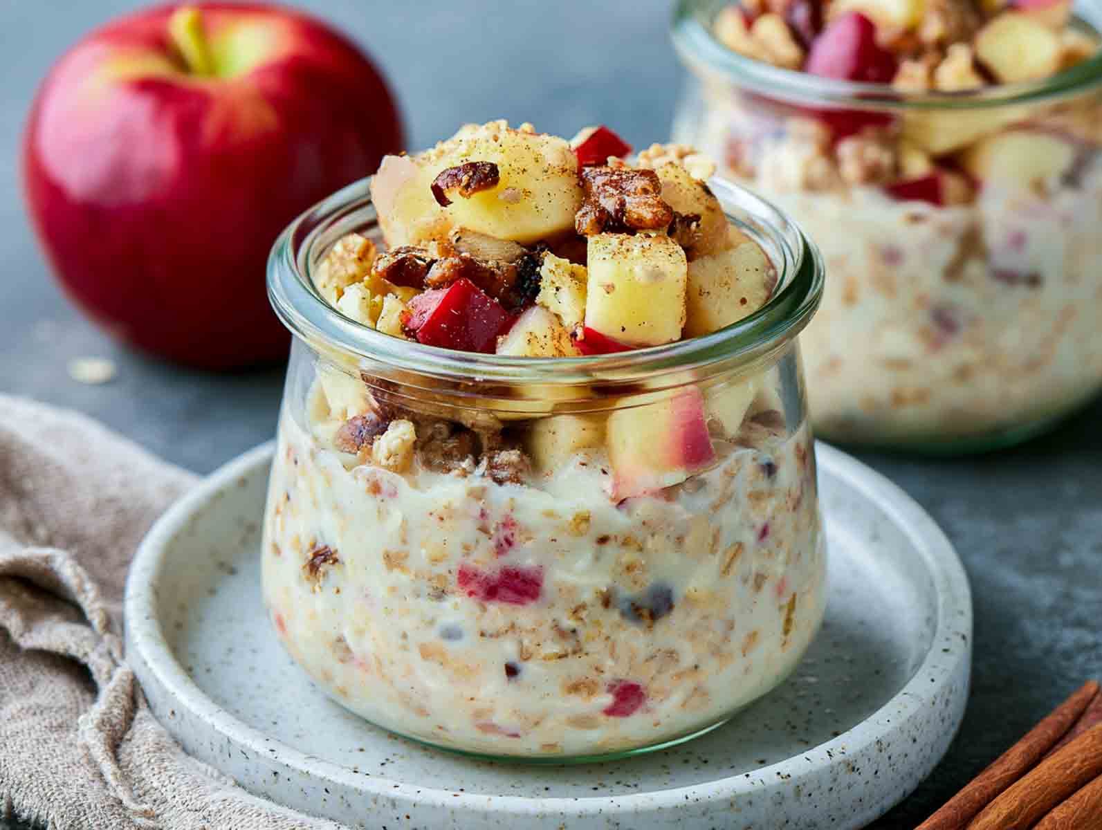 Apple pie kefir overnight oats topped with diced red apple, pecans, and cinnamon, with a whole apple behind.