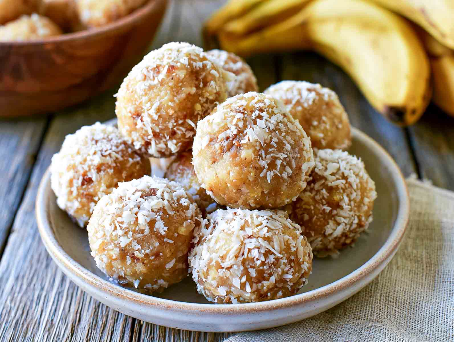 Banana coconut high-protein energy balls rolled in shredded coconut on a ceramic plate, with ripe bananas behind.