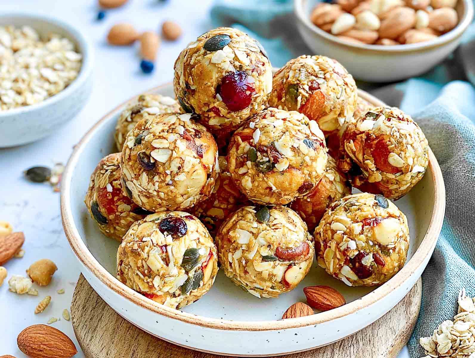 Breakfast granola high-protein energy balls packed with oats, almonds, pepitas, and dried cranberries on a white plate, with bowls of oats and almonds.