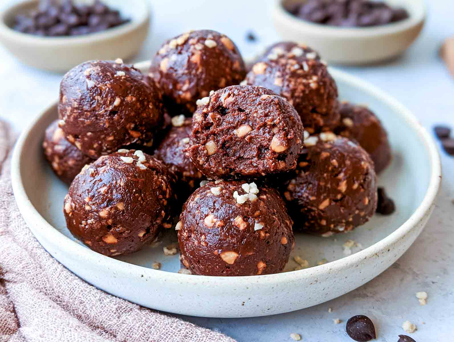 No-bake brownie batter high-protein energy balls studded with chopped peanuts, one bitten to show the fudgy inside, with bowls of chocolate chips behind.