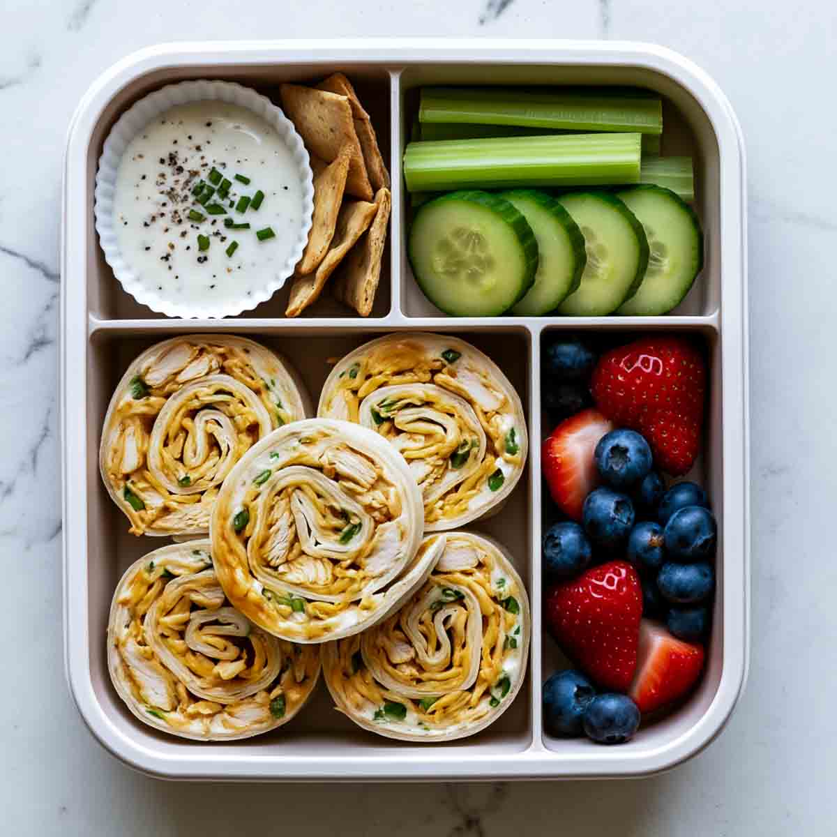 Bento lunchbox filled with buffalo ranch chicken pinwheels, ranch dip with crackers, cucumber and celery sticks, and fresh strawberries and blueberries.