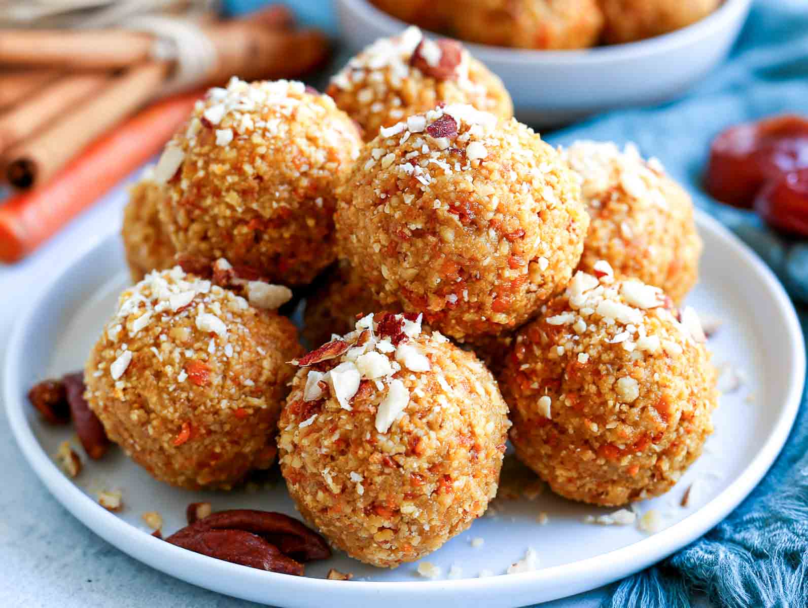 Carrot cake high-protein energy balls flecked with grated carrot and topped with chopped almonds, with cinnamon sticks, pecans, and dried apricots nearby.