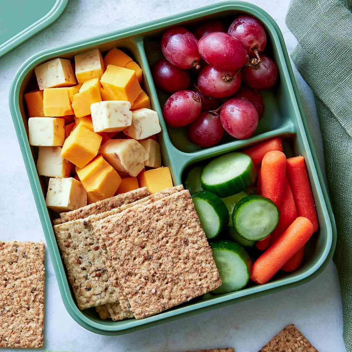 Cheese cubes, whole-grain crackers, grapes, cucumbers, and baby carrots packed in a lunchbox.