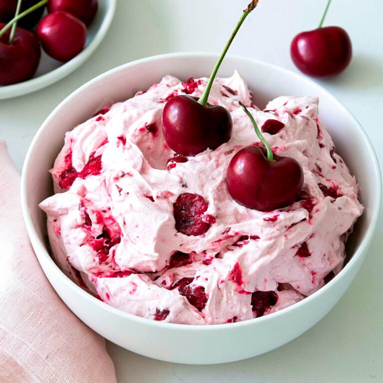 Cherry cheesecake fluff in a white bowl, topped with fresh cherries, with a pink napkin and extra cherries in the background.