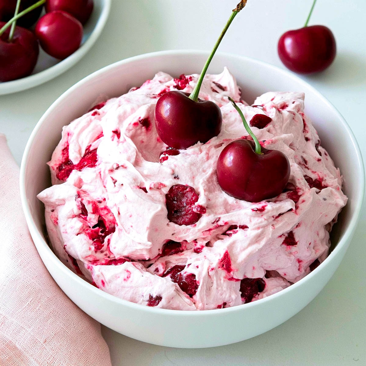 Cherry cheesecake fluff in a white bowl, topped with fresh cherries, with a pink napkin and extra cherries in the background.