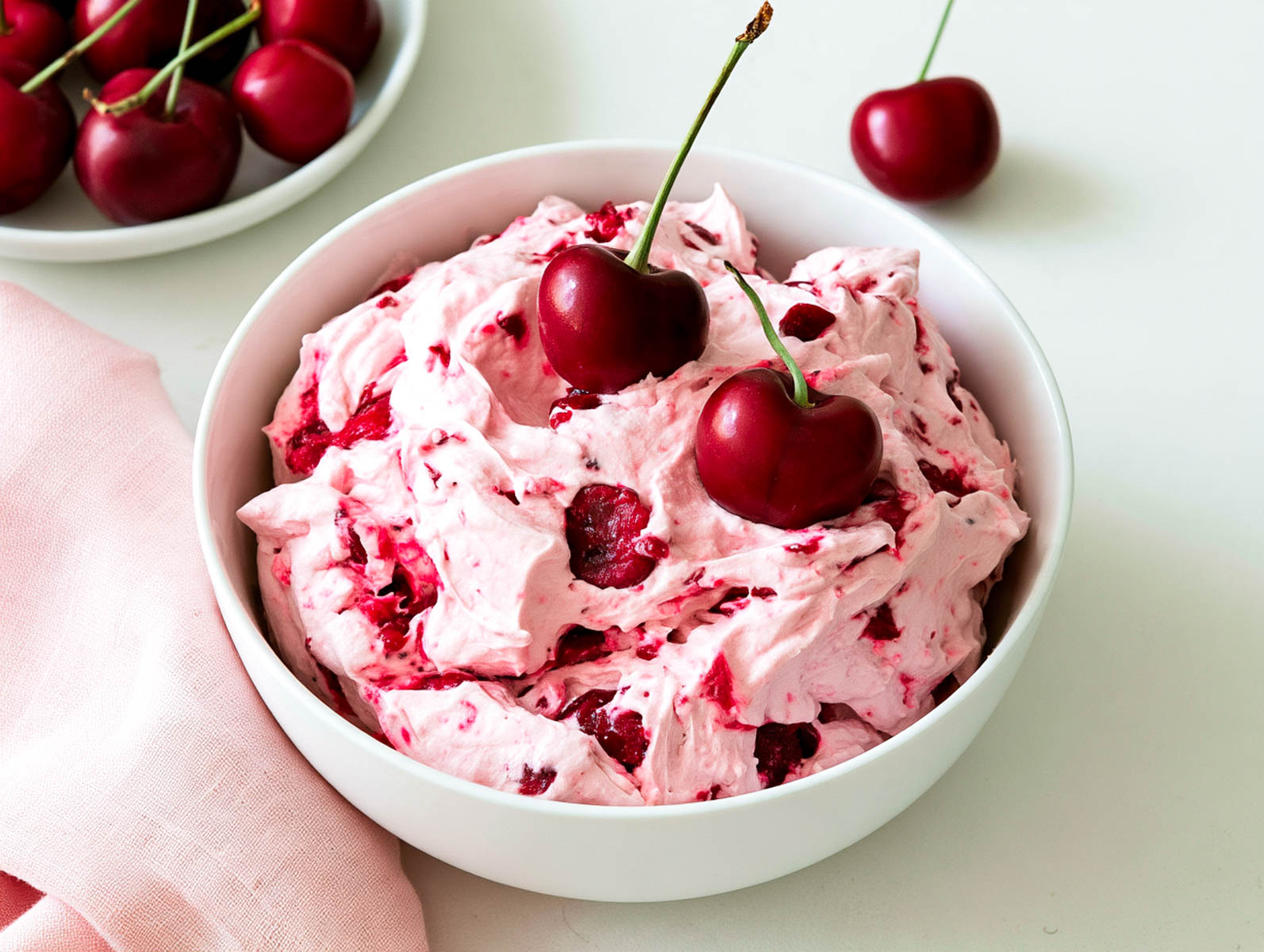 Close-up of creamy pink cherry cheesecake fluff in a white bowl, studded with cherries and topped with two fresh cherries.