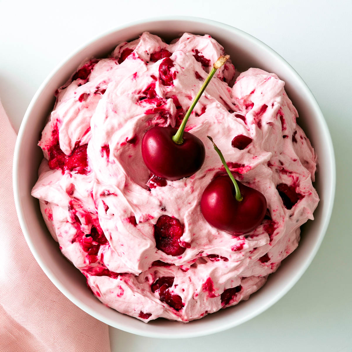 Top-down view of cherry cheesecake fluff with visible cherry pieces and two fresh cherries on top, served in a white bowl.