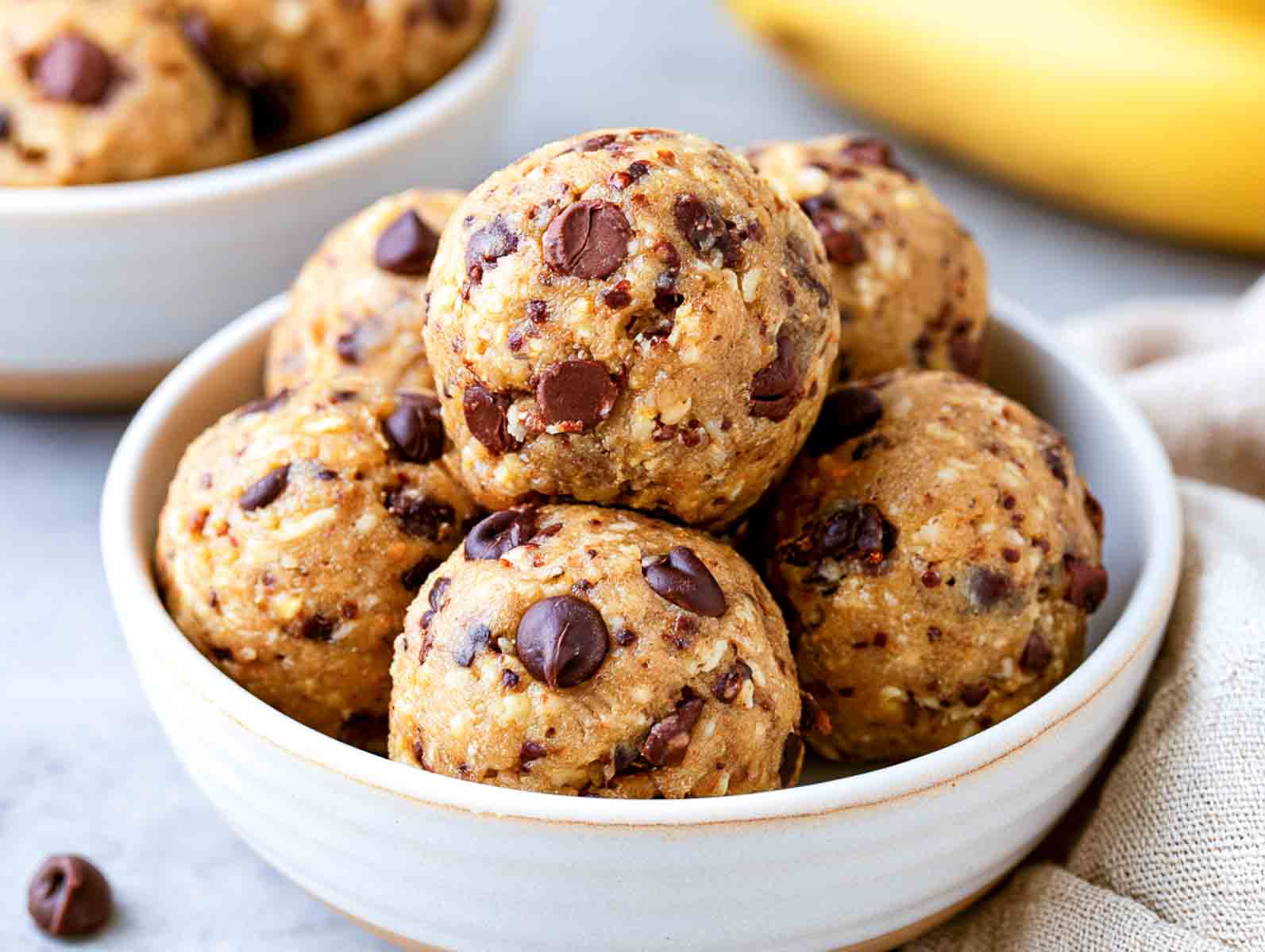 Chocolate chip banana high-protein breakfast energy balls studded with chocolate chips in a white bowl, with a banana behind.