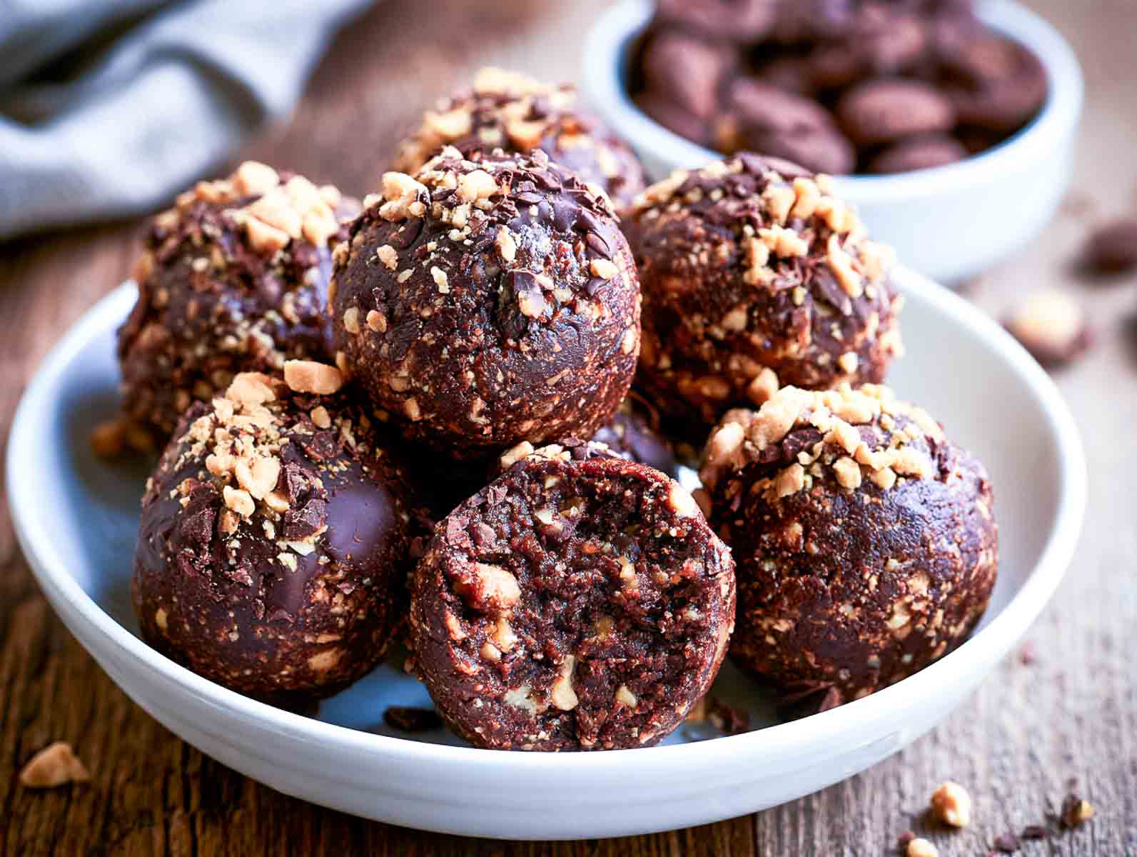 Keto chocolate peanut butter high-protein energy balls studded with chopped peanuts, one bitten to show the fudgy inside, with bowls of chocolate chips behind.
