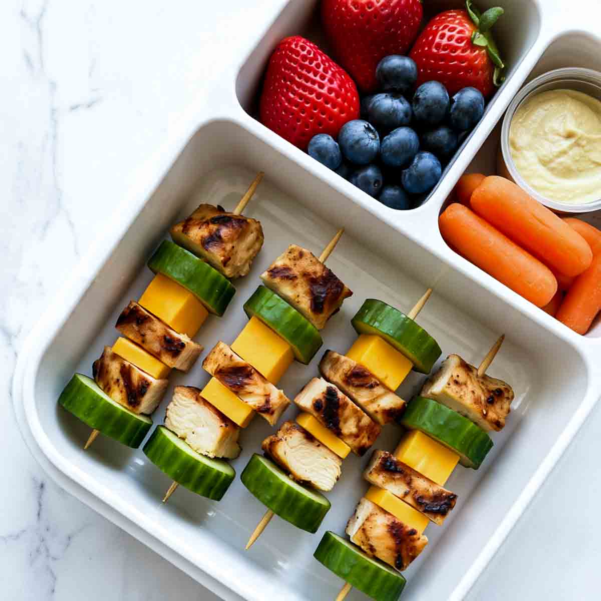 Lunchbox with mini chicken, cheddar cheese, and cucumber skewers, served with strawberries, blueberries, carrot sticks, and a creamy dip.