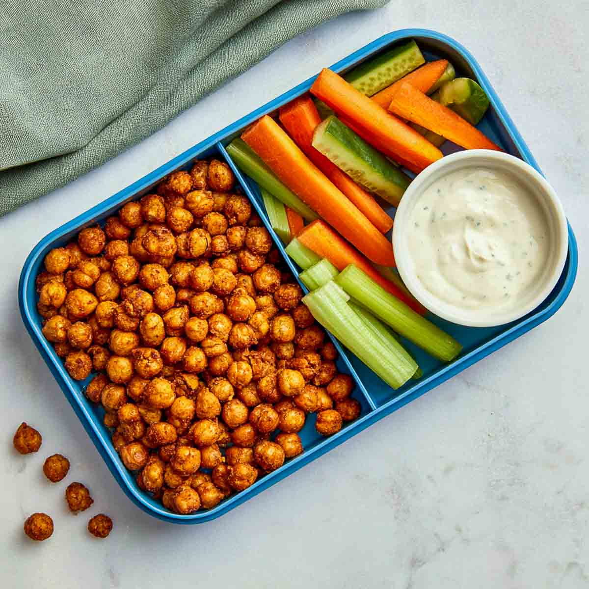 Crispy roasted chickpeas with ranch dip, cucumber, celery, and carrots in a blue lunchbox.