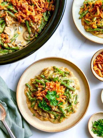 Crockpot green bean casserole on a stoneware plate topped with crispy fried onions and parsley, beside the slow cooker.