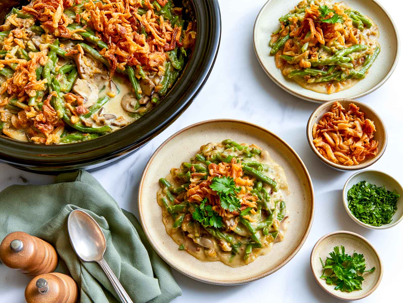 Crockpot green bean casserole with sautéed mushrooms and crispy fried onions in a black slow cooker, served with a plate of casserole on a white marble surface.