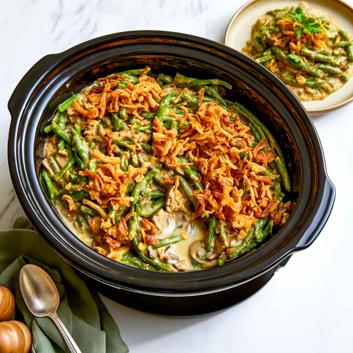 Black slow cooker filled with creamy green bean casserole, sliced mushrooms, and crispy fried onions, photographed from above on a white marble countertop.