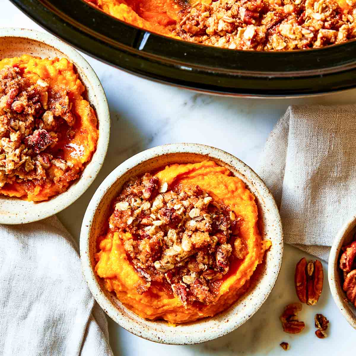 Two speckled bowls of mashed sweet potato casserole with crunchy pecan oat streusel next to a slow cooker.